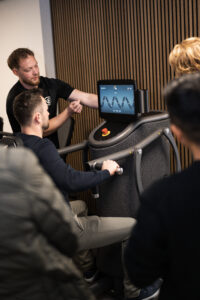 Rens Steenbergen als fitcoach van Vita Fortis op de foto. Hij begeleid een man op de Chest Press bij Vita Fortis. Er staan meerdere toeschouwers bij