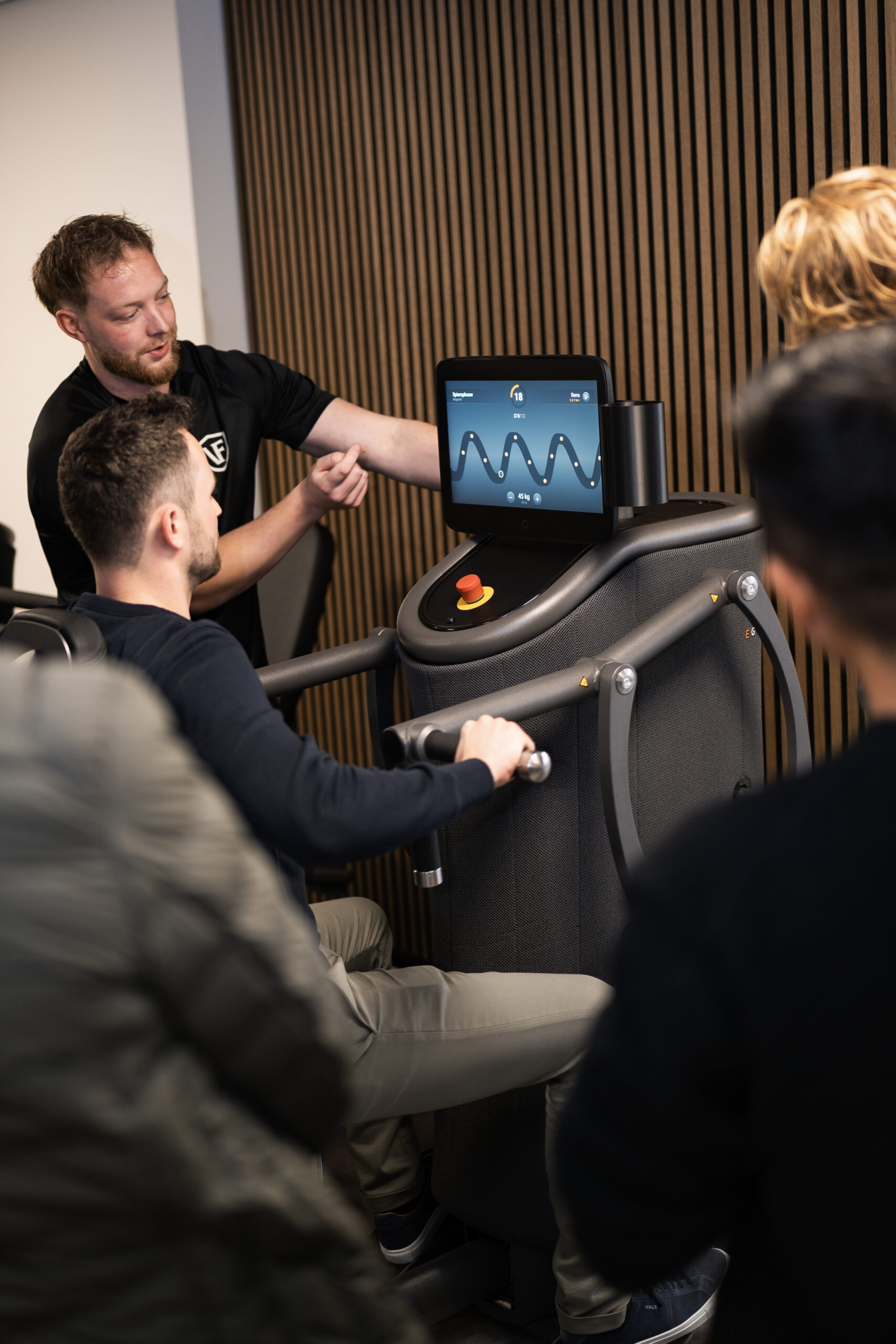 Rens Steenbergen als fitcoach van Vita Fortis op de foto. Hij begeleid een man op de Chest Press bij Vita Fortis. Er staan meerdere toeschouwers bij